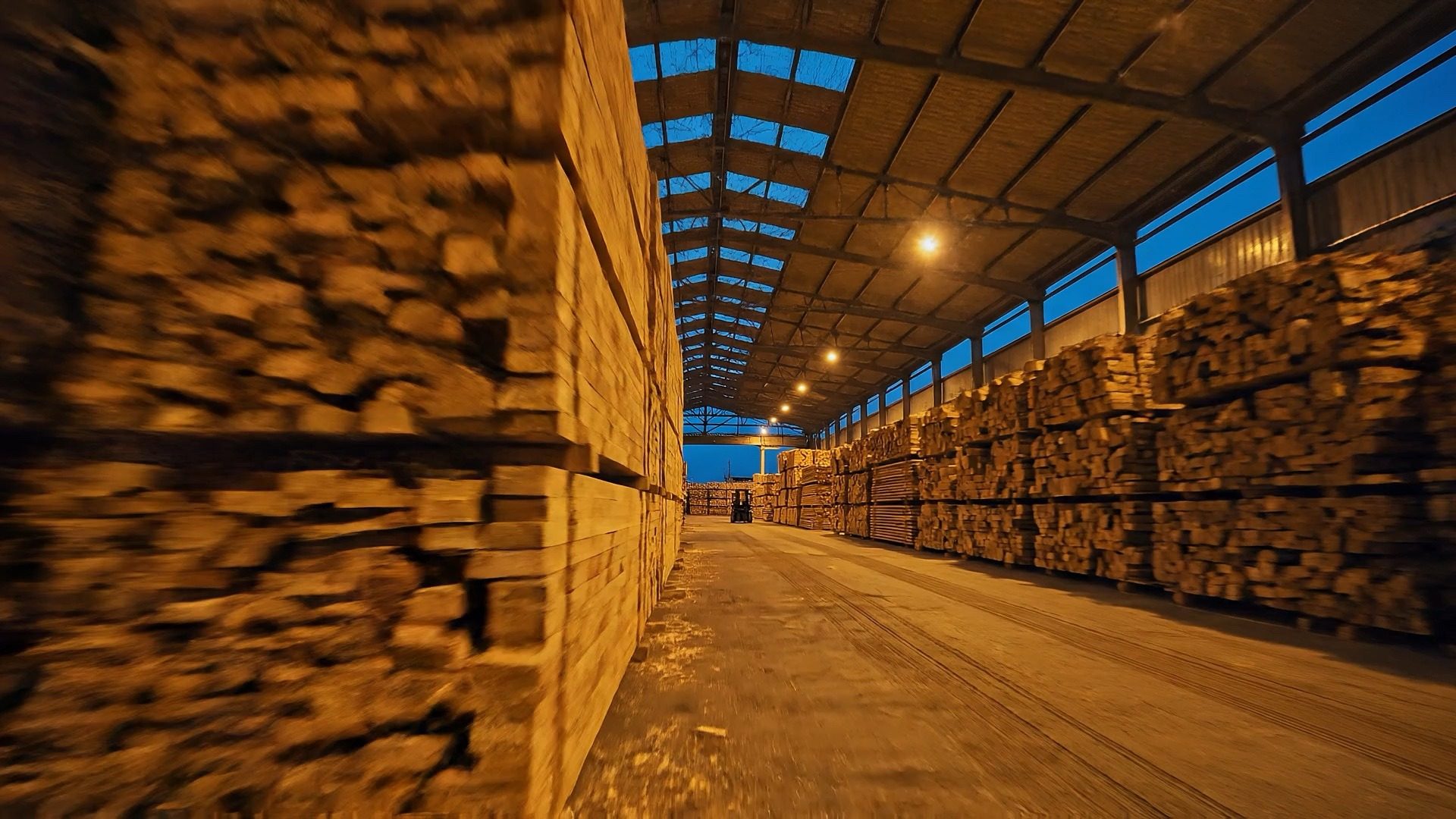Stacks of sawn timber in a covered timber yard at dusk, a forklift at the end of the aisle