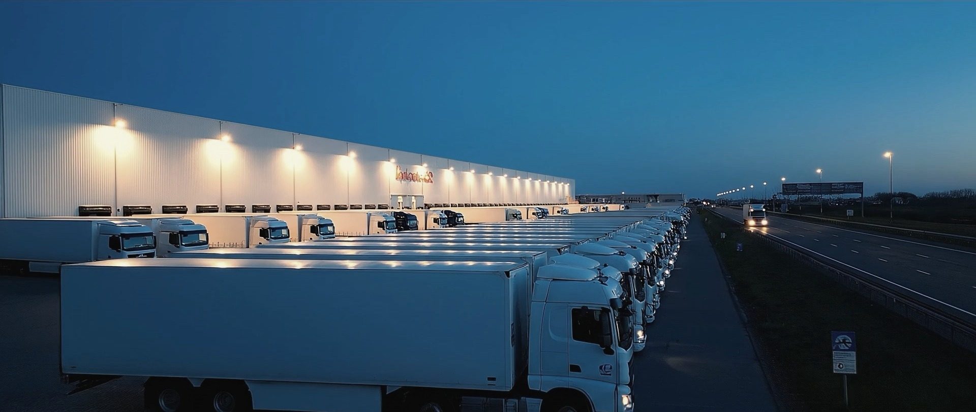 A long row of lorries parked along a lit distribution centre at dusk, with the motorway running past