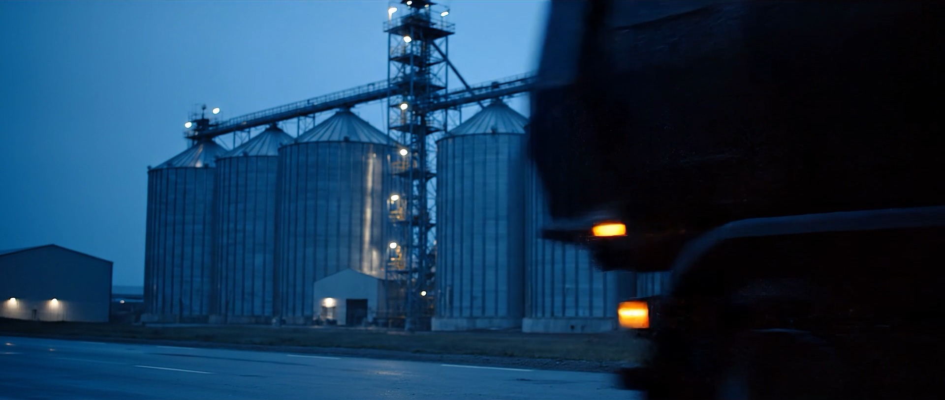 Truck driving past floodlit grain silos at dusk
