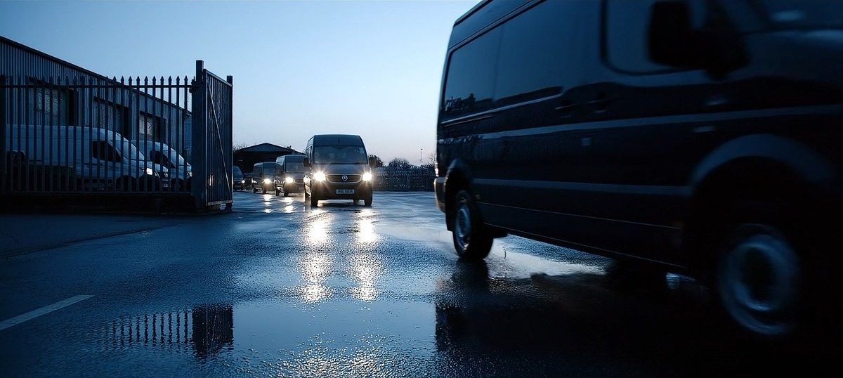 Delivery vans with headlights on driving out of a depot gate at dusk